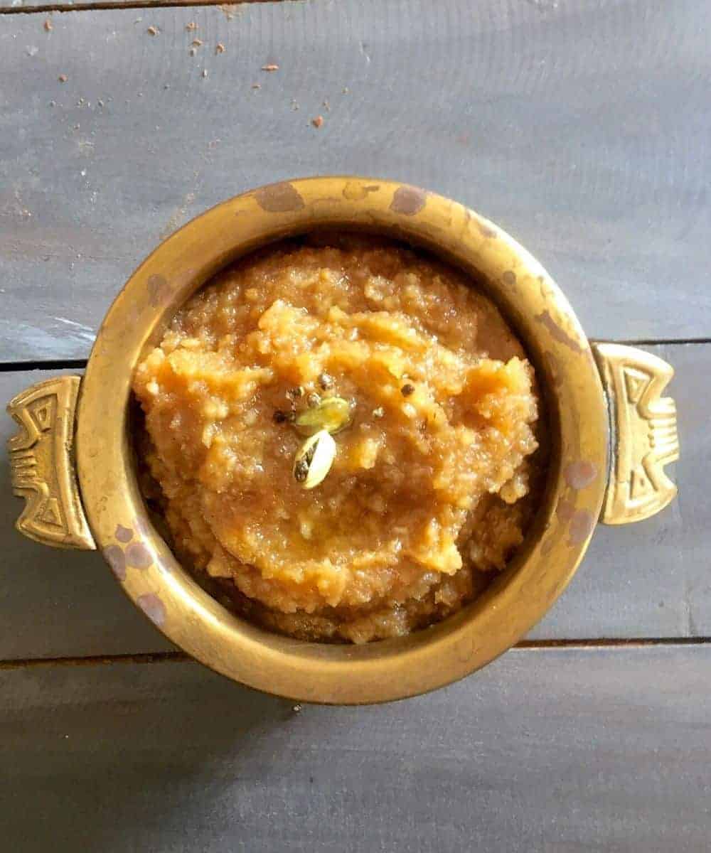 Golden brown kali, a sweet rice and jaggery dish with cardamom pods garnish, in a brass two handled bowl, on a wooden background