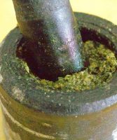 A stone mortar with pestle partly filled with fresh ground basil pesto. On a yellow background