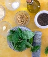 Ingredients for making pesto: A stone mortar with basil leaves, the pestle by its side; a glass bowl with roasted pine nuts, another with 2 cloves of garlic, a small mortar with groud black pepper, a bottle of olive oil, a bowl of grated parmesan and another of salt