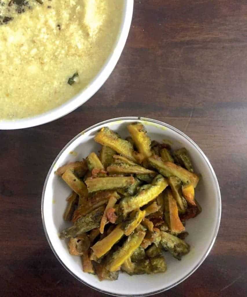 Fried strips of bitter gourd/ karela in a white bowl with yogurt based yellow morekuzhambu in another white bowl diagonally above, all on a brown wooden background
