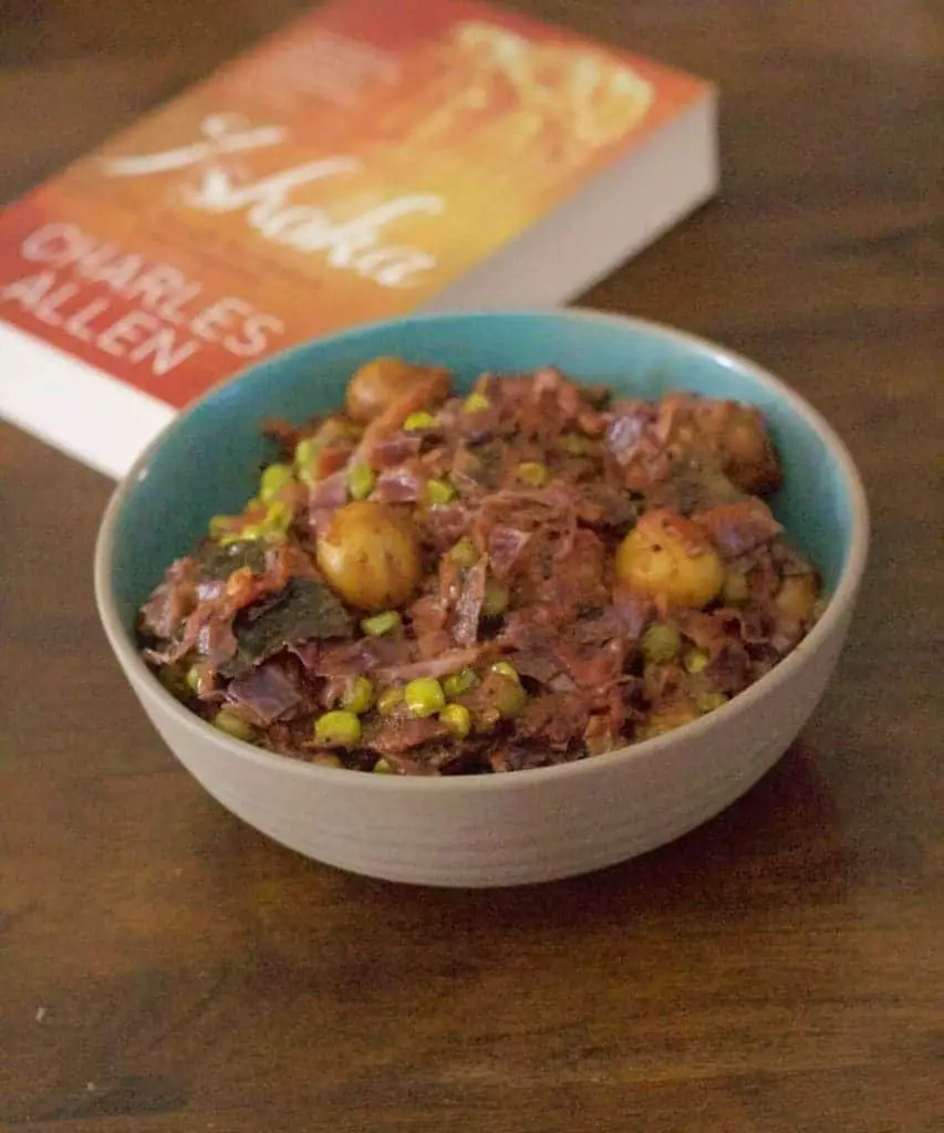 Red cabbage strips cooked with baby potatoes and green peas cooked with mild spices, in a blue bowl brown on the outside. A book on Indian emperor ashoka behind the bowl, all on a brown wooden background