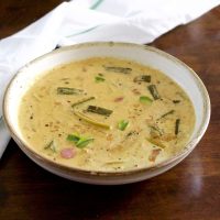 Pale yellow creamy raw mango curry with okra, shallots, mango and curry leaves floating on top, in a white bowl edged with beige. On a dark brown background with a white napkin with green stripe behind
