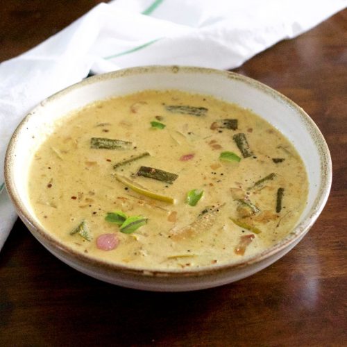 Pale yellow creamy raw mango curry with okra, shallots, mango and curry leaves floating on top, in a white bowl edged with beige. On a dark brown background with a white napkin with green stripe behind