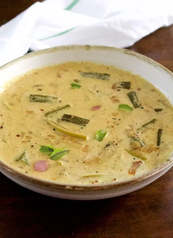 Pale yellow creamy raw mango curry with okra, shallots, mango and curry leaves floating on top, in a white bowl edged with beige. On a dark brown background with a white napkin with green stripe behind