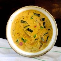 Pale yellow creamy raw mango curry with okra, with bits of okra and pink shallots floating on the surface. On a black background with a white napkin with narrow green stripes in the foreground