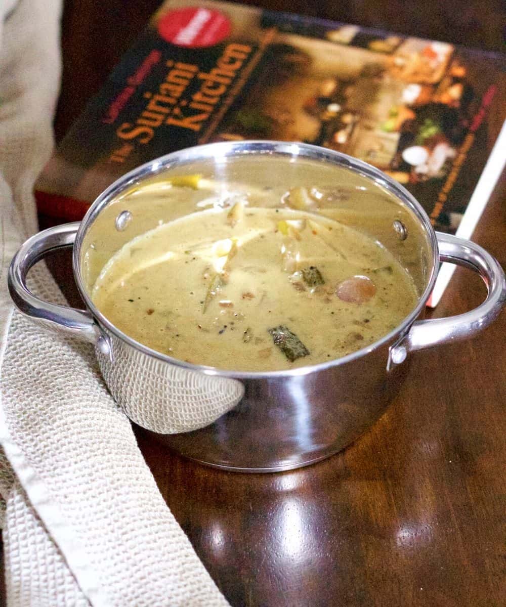 Steel bowl with 2 handles half filled with a cremy stew on which shallots, mango slices and okra slices can be seen. A beige napkin to the left and a brown and orange headed cook book to the right, all on a dark brown background