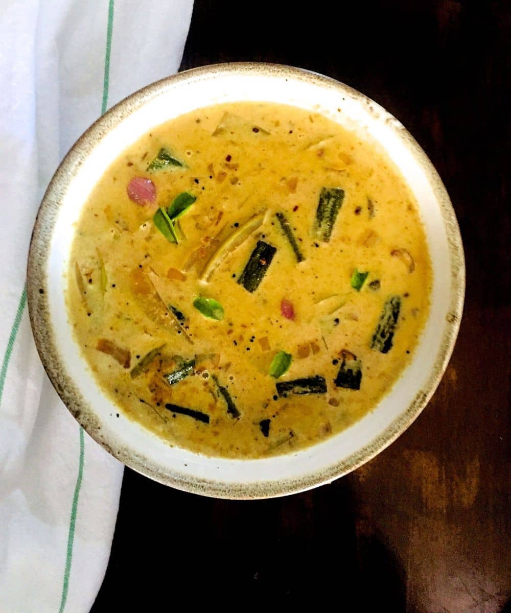 Yellow mustardy coloured stew with pieces of bhindi/ okra, green mango and pink shallots floating, in a white bowl edged with beige. All on a dark brown background and a white napkin with a thin green stripe on the side