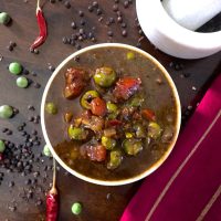 Brown pachai sundakkai puli kuzhambu in a bowl with green turkey berry and red tomatoes heaped on top. Peppercorns, red chilies, turkey berry scattered around a white mortar pestle. Red silk fabric to one side