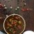 Overhead view of bowl of browntamarind and spice based gravy with green turkey berries, red tomato on top. Dry chili, peppercorn and green berries scattered around on brown background. White mortar pestle at edge of image