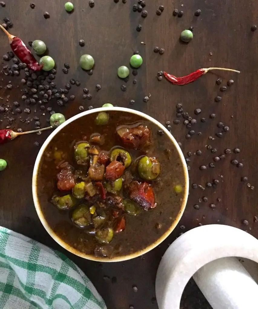 Dark brown tamarind based gravy with green turkey berries/ sundakkai, red tomato pieces on top. Peppercorns, green berries, red chilies scattered on brown backrground