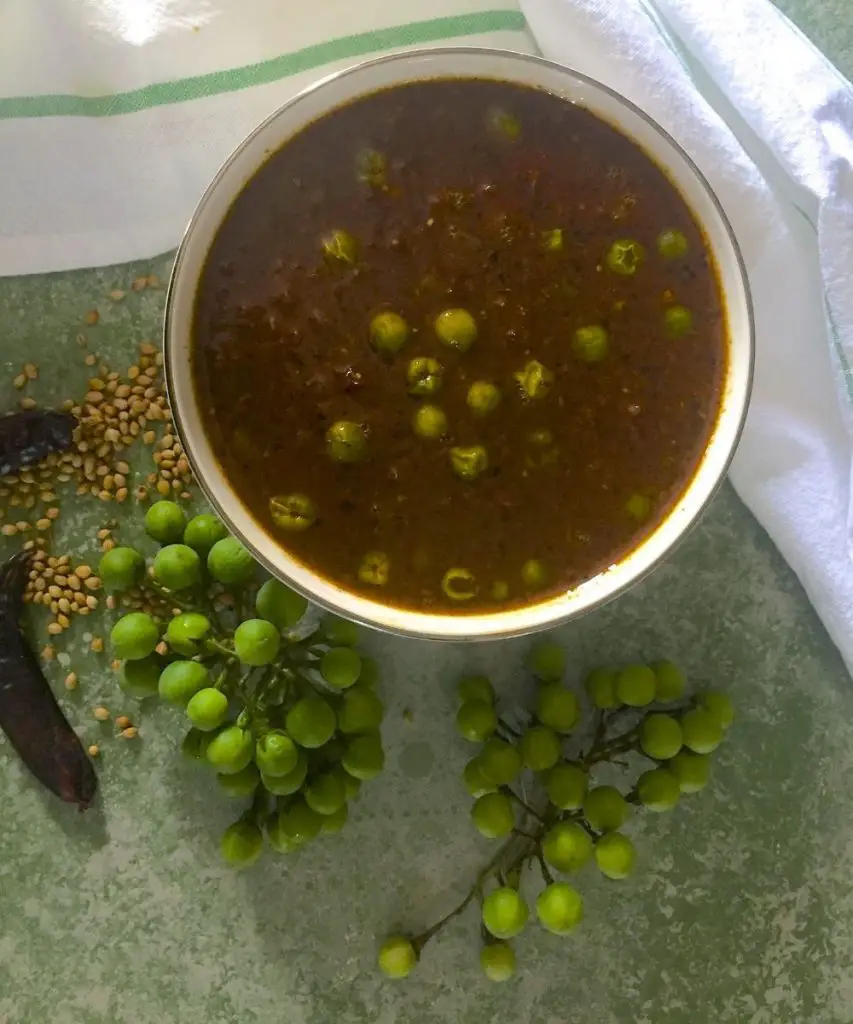 Bowl of brown tamarind based gravy with spices and spice paste with green cooked turkey berries/ sundakkai floating on top. Bunches of berries, some spices and dry chili to the left, all on a green background.