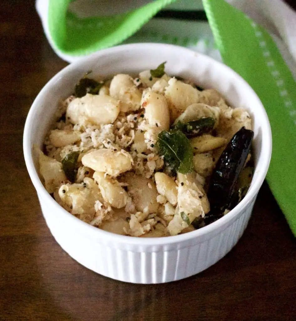 Cream coloured cooked mochai double beans in a white bowl with grated coconut and a dry red chili, curry leaves tempering. A green and white napkin to the left. All on a brown background