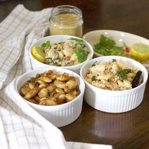 Three bowls each with a different flavour of cooked mochai double beans, one brown spiced, one with curry leaves, one with green coriander leaves. A beige spice mix jar in the back, a bowl with yellow lemon, red chili and green cilantro and a beige and white napkin to the left