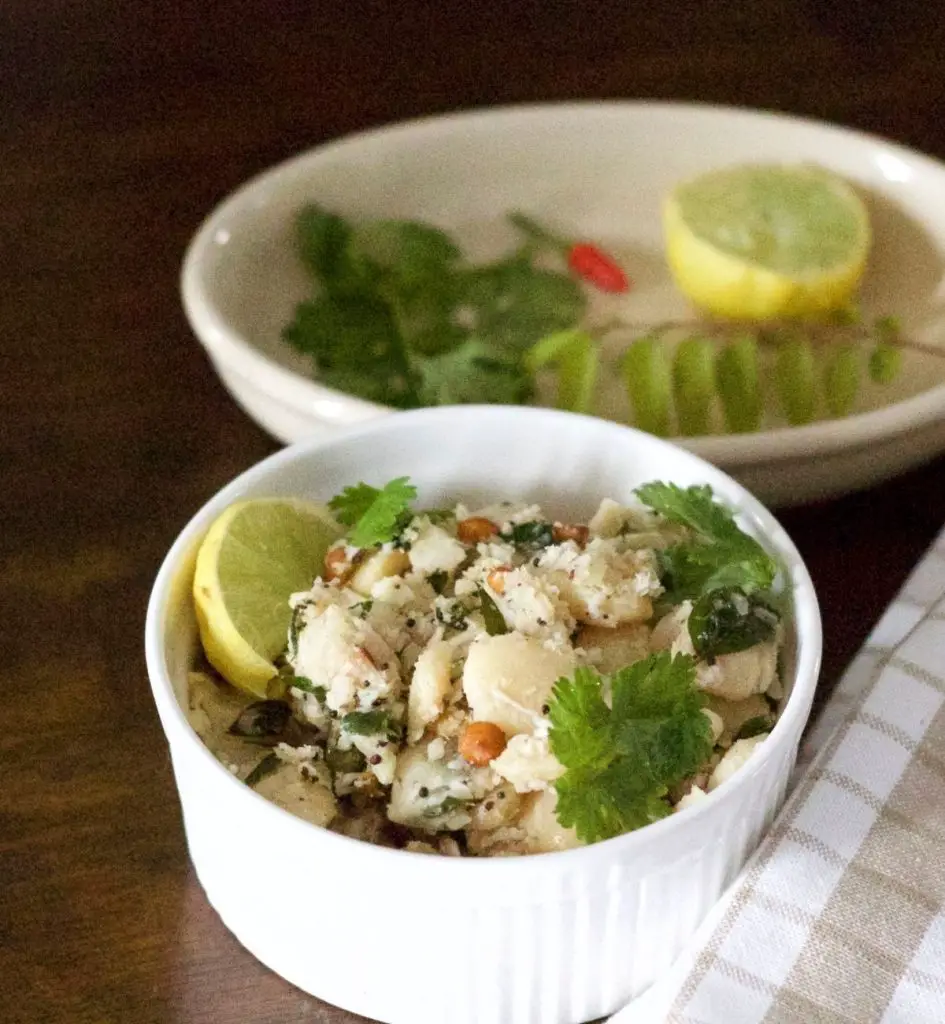 White bowl filled with white double beans mochai Sundal with tempering of channa dal, mustard, garnished with coconut and coriander leaves and a slice of lemon at one side. A beige and white napkin to the right and a plate with lemon, red chili and curry leaves, coriander leaves at the back