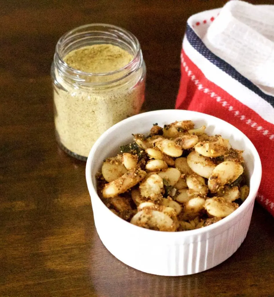 White bowl filled with double beans mochai Sundal in a homemade spice curry mix. A glass jar with beige coloured spice mix at the back, a red and white napkin on the right, all on a brown background