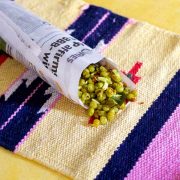 Cooked dry green peas with coconut raw mango and peanuts wrapped in a roll of newspaper resting on a sand and pink coloured mat on a yellow background