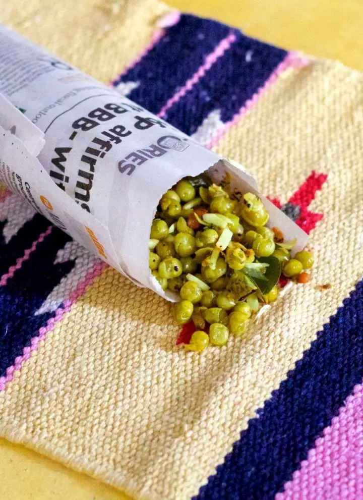 Cooked dry green peas with coconut raw mango and peanuts wrapped in a roll of newspaper resting on a sand and pink coloured mat on a yellow background