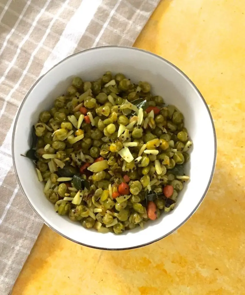 Thenga Manga Pattani Sundal. White bowl with cooked green peas, coconut and raw mango shavings and peanuts, on a yellow background with a beige and white checked napkin to the left