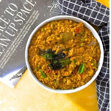 Bisi Bele Huli Anna, a Brownish yellow hued rice and lentils mix with mustard seeds and green curry leaves tempering, in a white bowl on a yellow background. Black page of a newspaper to the left and a napkin with black squares to the right.