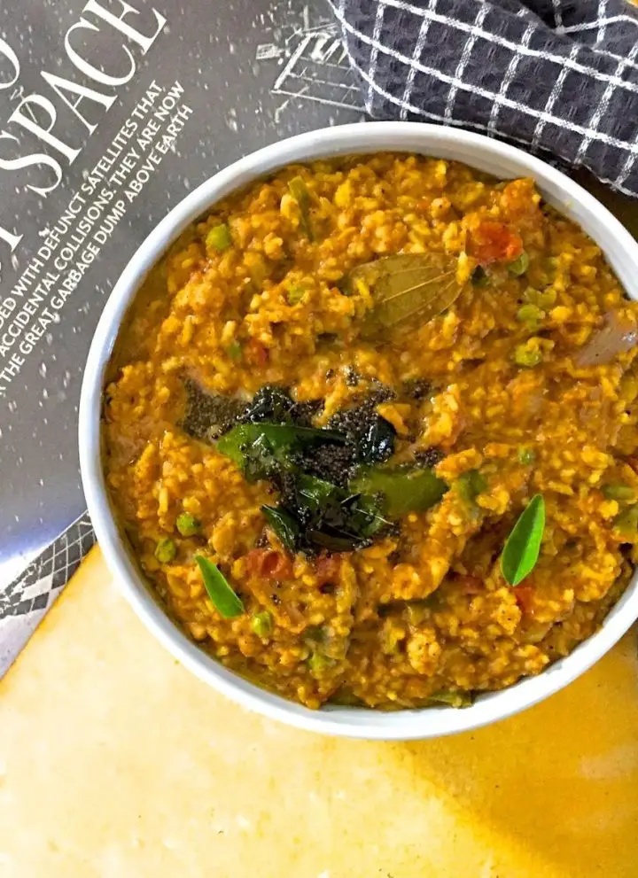 Bisi Bele Huli Anna, a Brownish yellow hued rice and lentils mix with mustard seeds and green curry leaves tempering, in a white bowl on a yellow background. Black page of a newspaper to the left and a napkin with black squares to the right.
