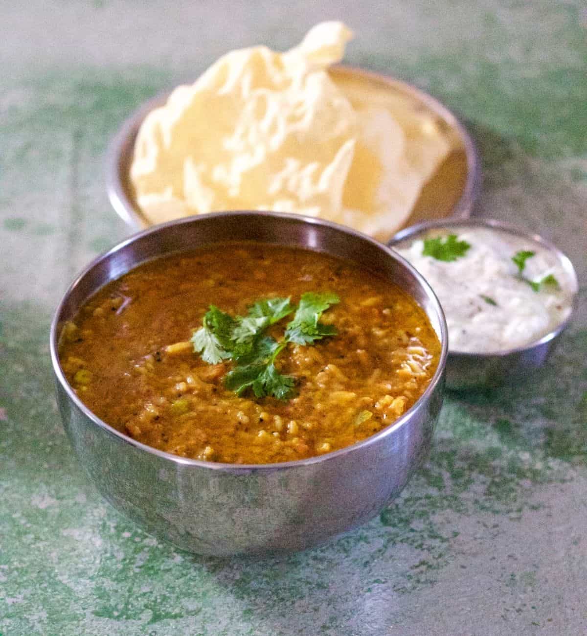 Large steel bowl filled with brownish yellow bisi bele huli anna, a rice and lentils spiced mix. Garnished with green coriander leaves. Plate of fried appalam/pappad in the background and a bowl of curd pachadi/ raita to the right. On a green and white background