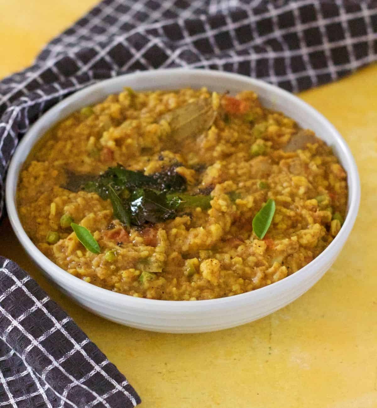 Wide bowl filled with a brown and yellow spiced rice (bisi bele huli anna) and lentil mix. Tempering of mustard seeds and curry leaves on top. Napkin with black squares edged with white in the background. All on a yellow background