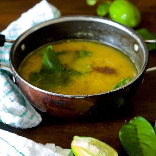 Pale yellow cooked lentils with gondhoraj lime leaves, green chili and radhuni spices seen on top. Green lime and leaves at background and foreground with slice of lime. Green and white napkin to the right. All on a brown wooden background