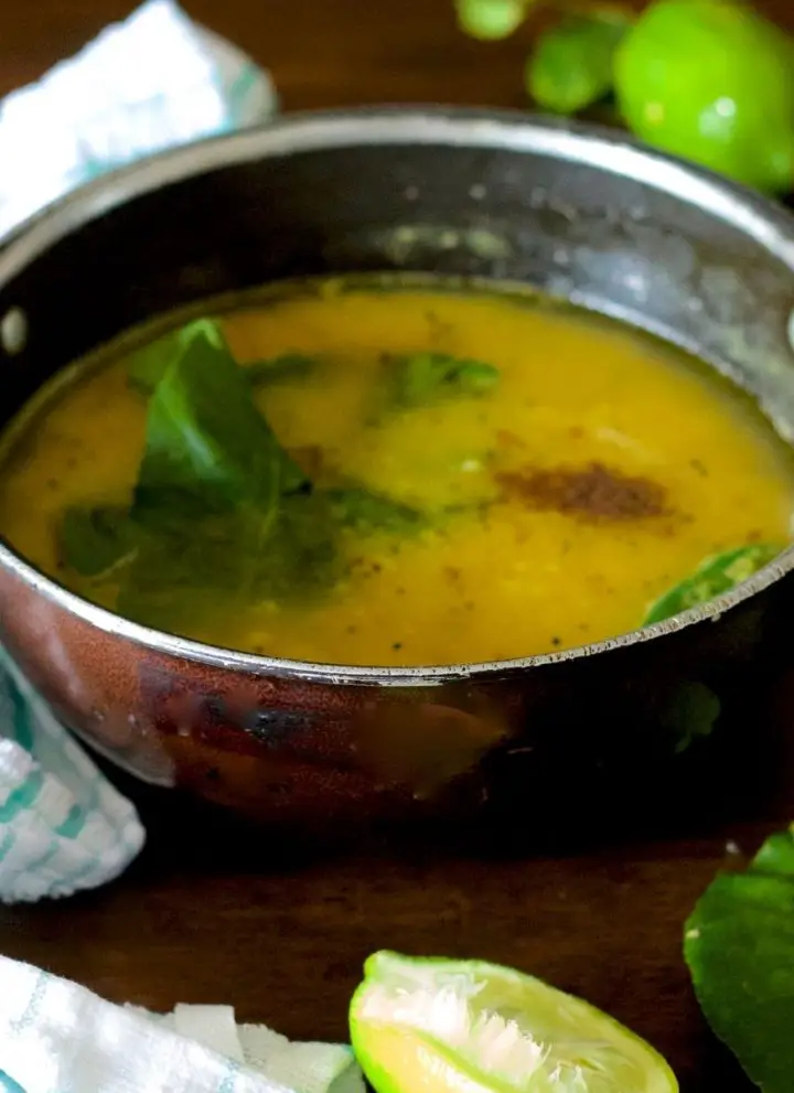 Pale yellow cooked lentils with gondhoraj lime leaves, green chili and radhuni spices seen on top. Green lime and leaves at background and foreground with slice of lime. Green and white napkin to the right. All on a brown wooden background