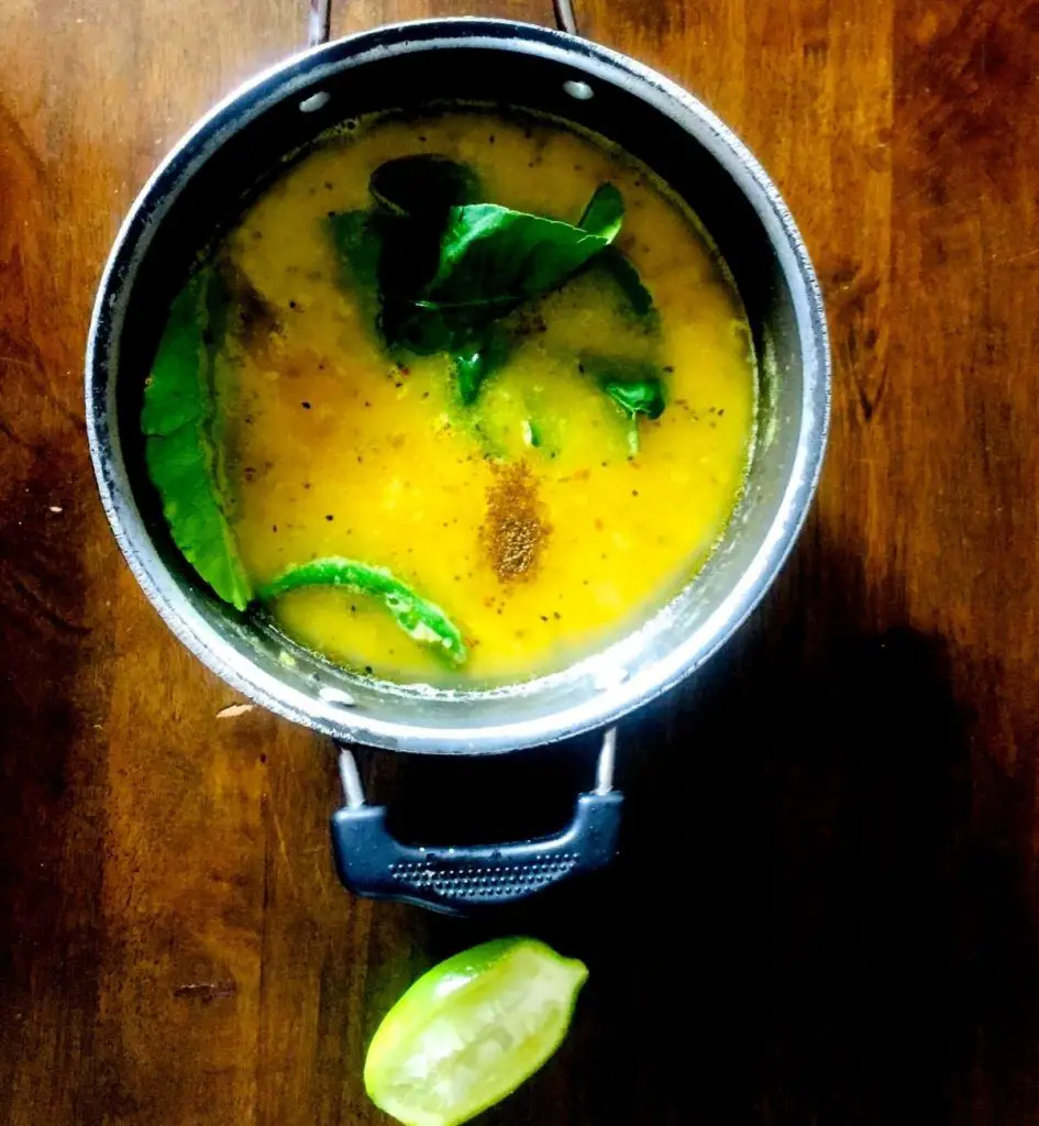 Gondhoraj Lebu Masoor Dal with Amada. Bright yellow cooked masoor lentils with green lime leaves, green chili and brown spices on the surface. Slice of lime in foreground. All on a brown background