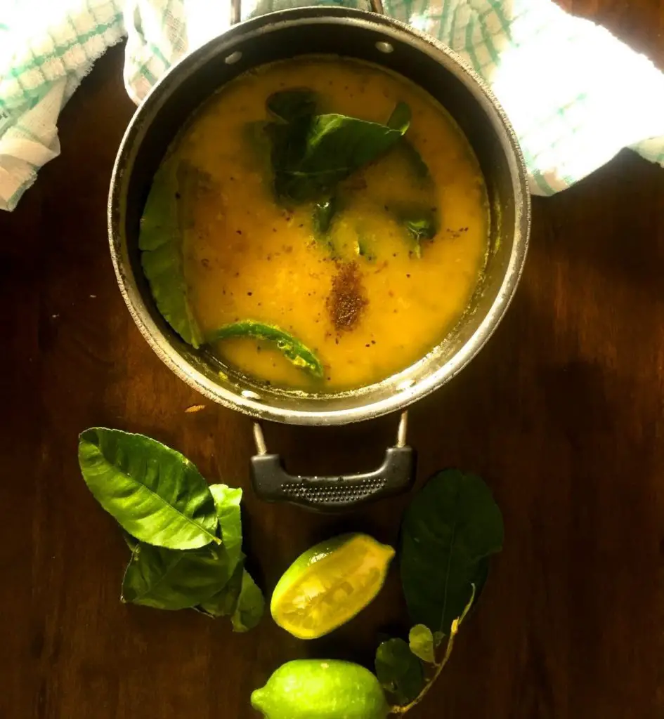 Pale yellow cooked lentils in a frying pan. Green chili, Lime leaves and brown spices on the surface. Lime leaves, sliced lime, whole green lime with stalk and leaves, all vertically below the pan.On a brown wooden background. Green and white napkin above in back ground