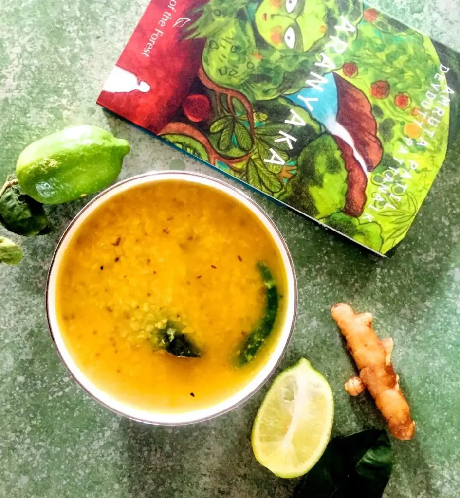 Bowl of yellow cooked masoor lentils dotted with brown spices, green chili and green lime leaf. Green Lime and leaves and a green covered book on Forests in the background, slice of lime and piece of brown mango ginger in the right foreground