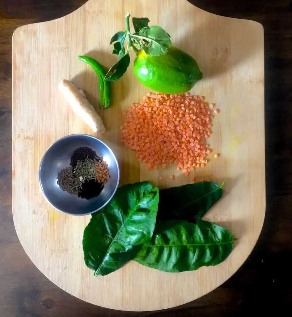 Ingredients for making Gondhoraj Lebu Masoor Dal, on a wooden cutting board. Vertically arranged, green lime with leaves and stalk; pink hued lentils, dark green lime leaves, stainless steel small bowl with brownish spices, stick of brown mango ginger