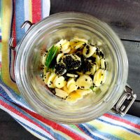 Pale yellow round slices of manga inji pickle in a glass jar. Bits of green chilies and black mustard seeds on top. An yellow, blue, white and red striped napkin to the left and front, all on a grey wooden board