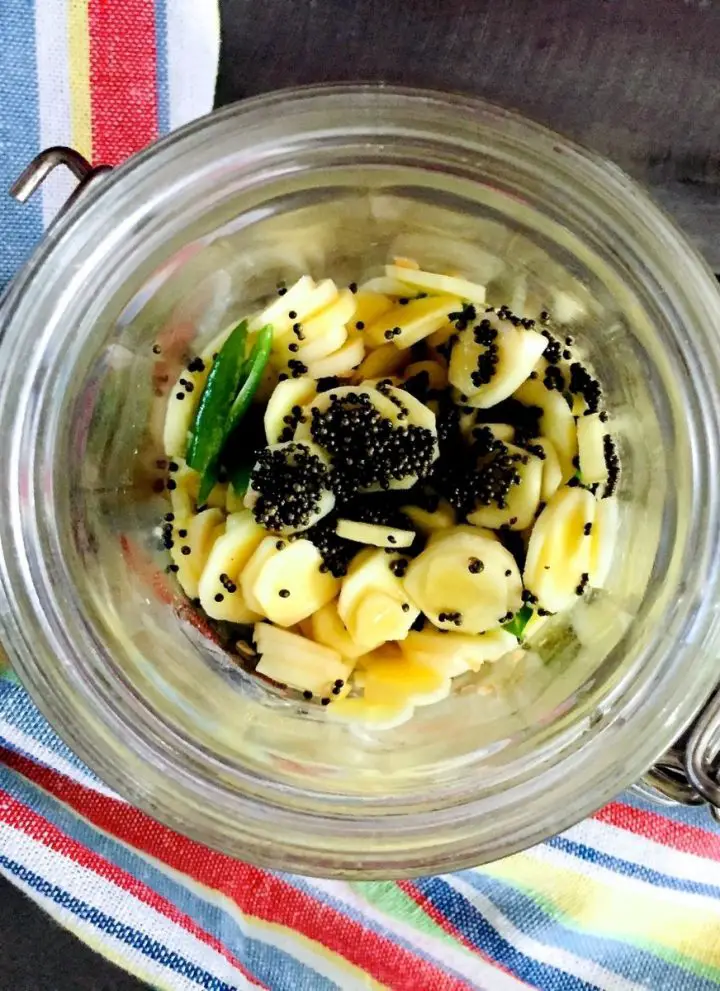 Pale yellow round slices of manga inji pickle in a glass jar. Bits of green chilies and black mustard seeds on top. An yellow, blue, white and red striped napkin to the left and front, all on a grey wooden board