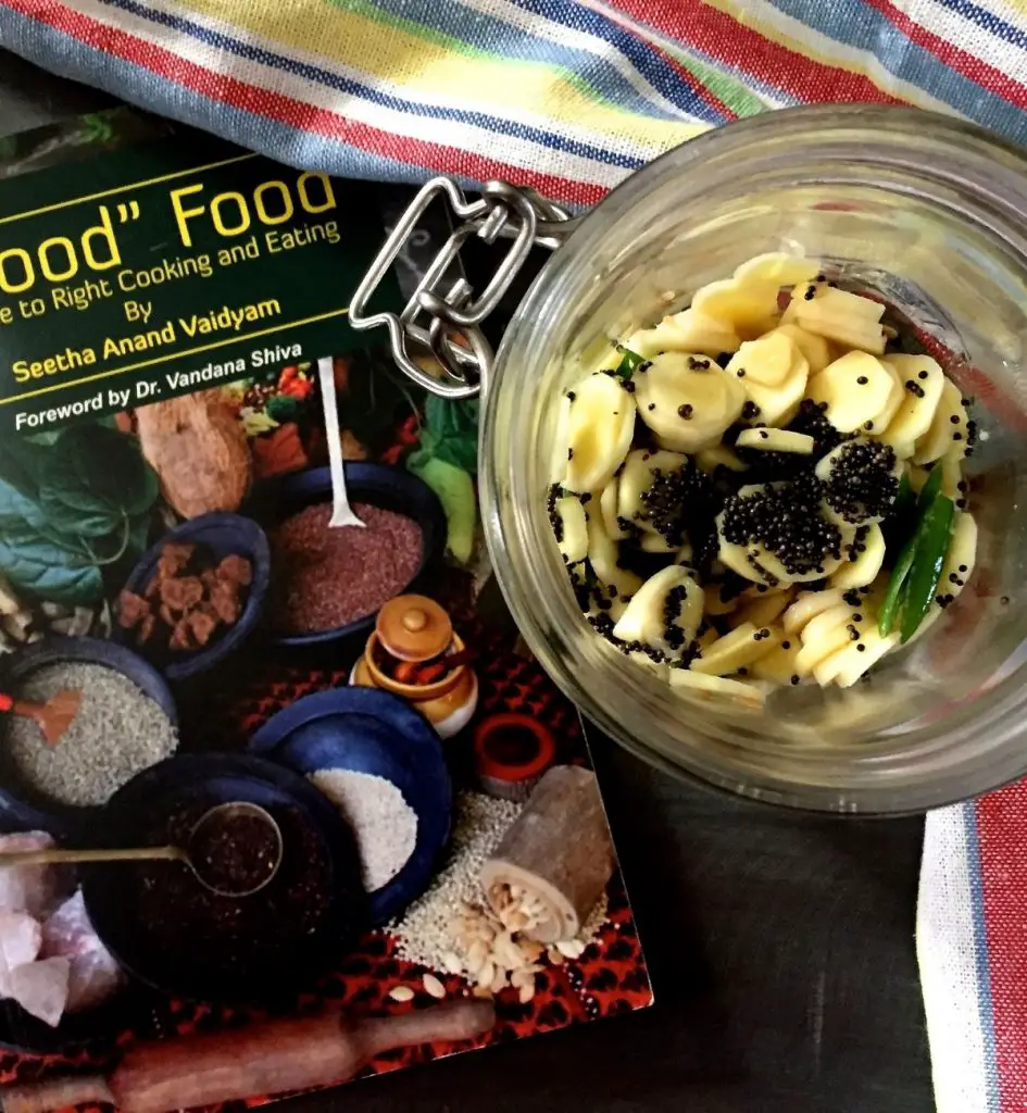 Yellow round slices of mango ginger with black mustard seeds and green chilli slices on top, in a glass jar seen from above. A dark green covered book with images of Indian spices, to the left. Multicoloured striped napkin of blue white red and yellow to the right