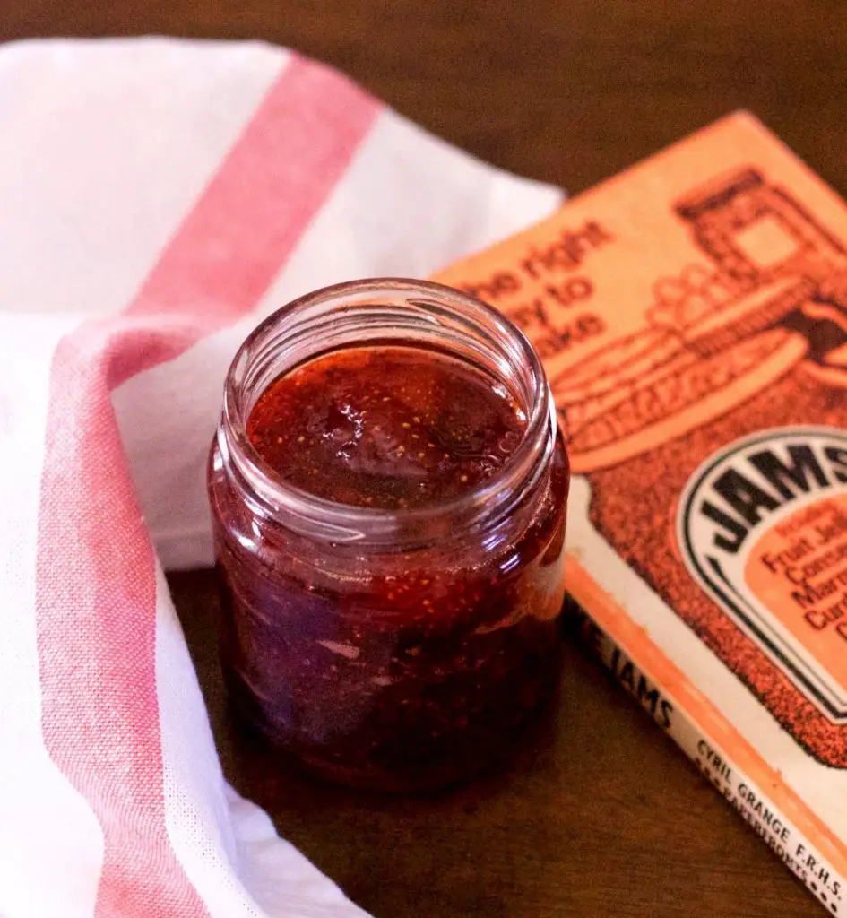 Lucious looking ruby red jam in a small glass jam jar. A pink and white striped napkin to the left and an orange coloured book on jam making to the right. On a brown wooden background