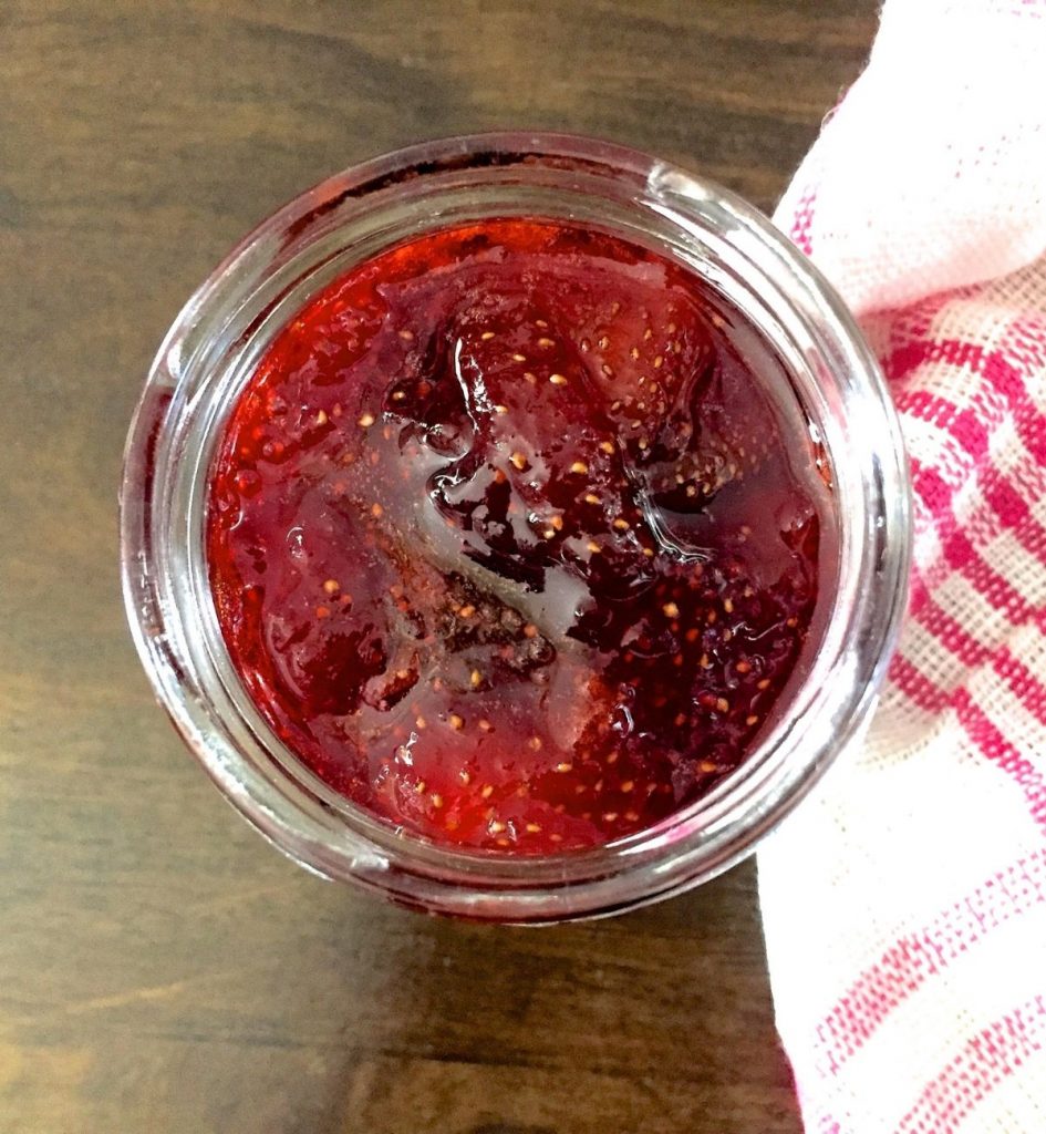Bright Ruby Red Jam in a glass jar, seen from above. Whole Strawberries can be seen. Pink and white napkin to the right. The whole on a plain brown wooden background