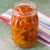 Tall glass jar with slices of vegetable pickle in oil, orange hued. Multicoloured striped napkin at the back. All on a green background