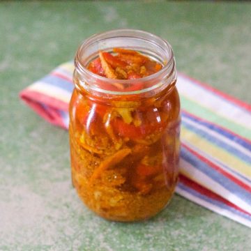 Gajar Gobi Shalgam Achar with Turmeric, orange hued pickle in a tall glass jar with slices of vegetables showing through. A green, blue, yellow, red, white striped napkin at the back, all on a dull green background