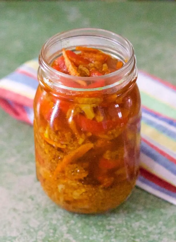 Gajar Gobi Shalgam Achar with Turmeric, orange hued pickle in a tall glass jar with slices of vegetables showing through. A green, blue, yellow, red, white striped napkin at the back, all on a dull green background