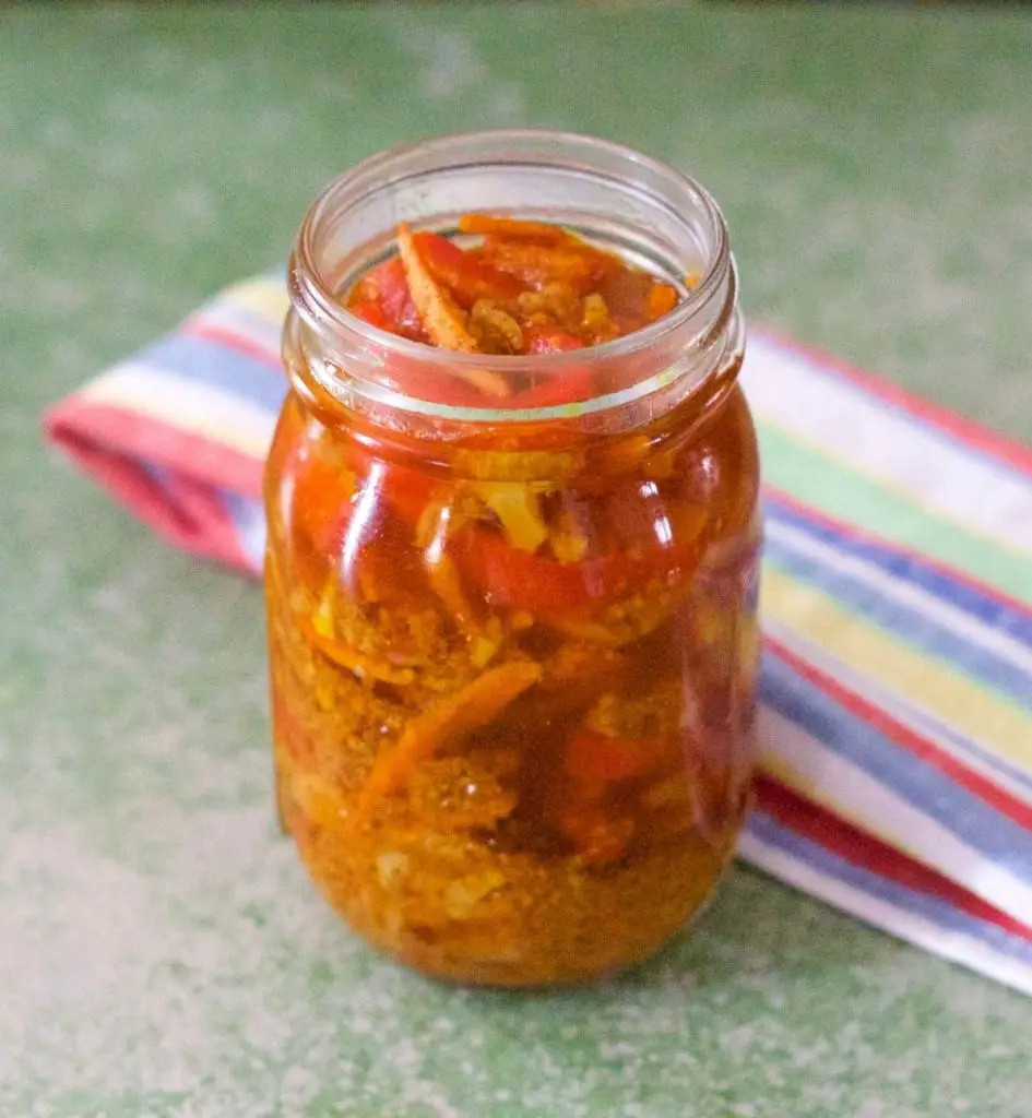 Gajar Gobi Shalgam Achar. Vegetables pickled in spices and mustard oil in a tall glass jar. Multicoloured striped napkin at the back, on a green background