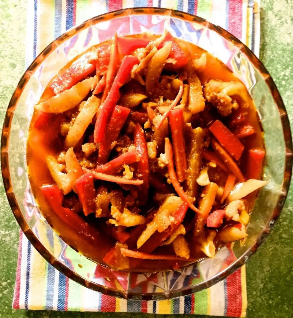 Slices of carrot, turnip, cauliflower, turmeric pickled in mustard oil with spices. Orange hued pickle in a glass bowl, on a red, yellow, green, blue, white striped napkin, on a green background