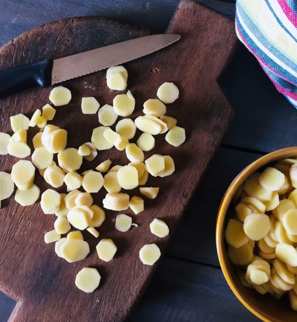 Several round slices of mango ginger/ manga inji/ amada/ amba haldi on a brown chopping board with a black handled knife. A bowl with more slices on the right and a napkin multi coloured with yellow, blue, red and white to the right background