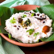 Clay bowl with a green banana leaf filled with white curd and rice, with garnish of green cilantro leaves and tempering of chilies and mustard seeds in oil, with a mango pickle seen on one side. A pale green napkin to the left