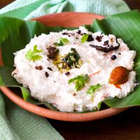 Clay bowl with a green banana leaf filled with white curd and rice, with garnish of green cilantro leaves and tempering of chilies and mustard seeds in oil, with a mango pickle seen on one side. A pale green napkin to the left