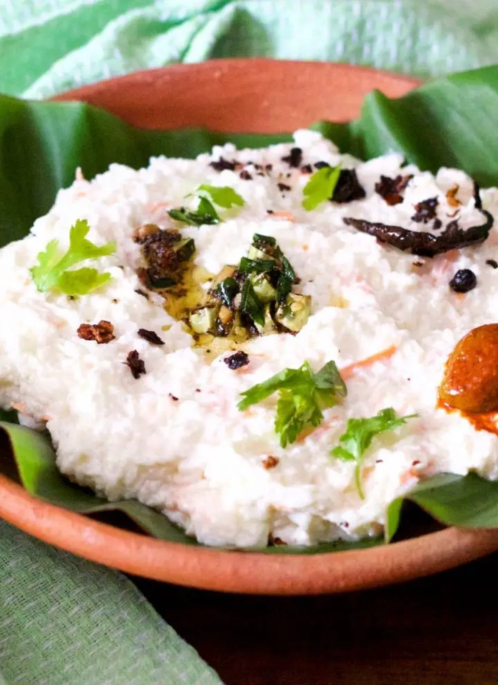 Clay bowl with a green banana leaf filled with white curd and rice, with garnish of green cilantro leaves and tempering of chilies and mustard seeds in oil, with a mango pickle seen on one side. A pale green napkin to the left