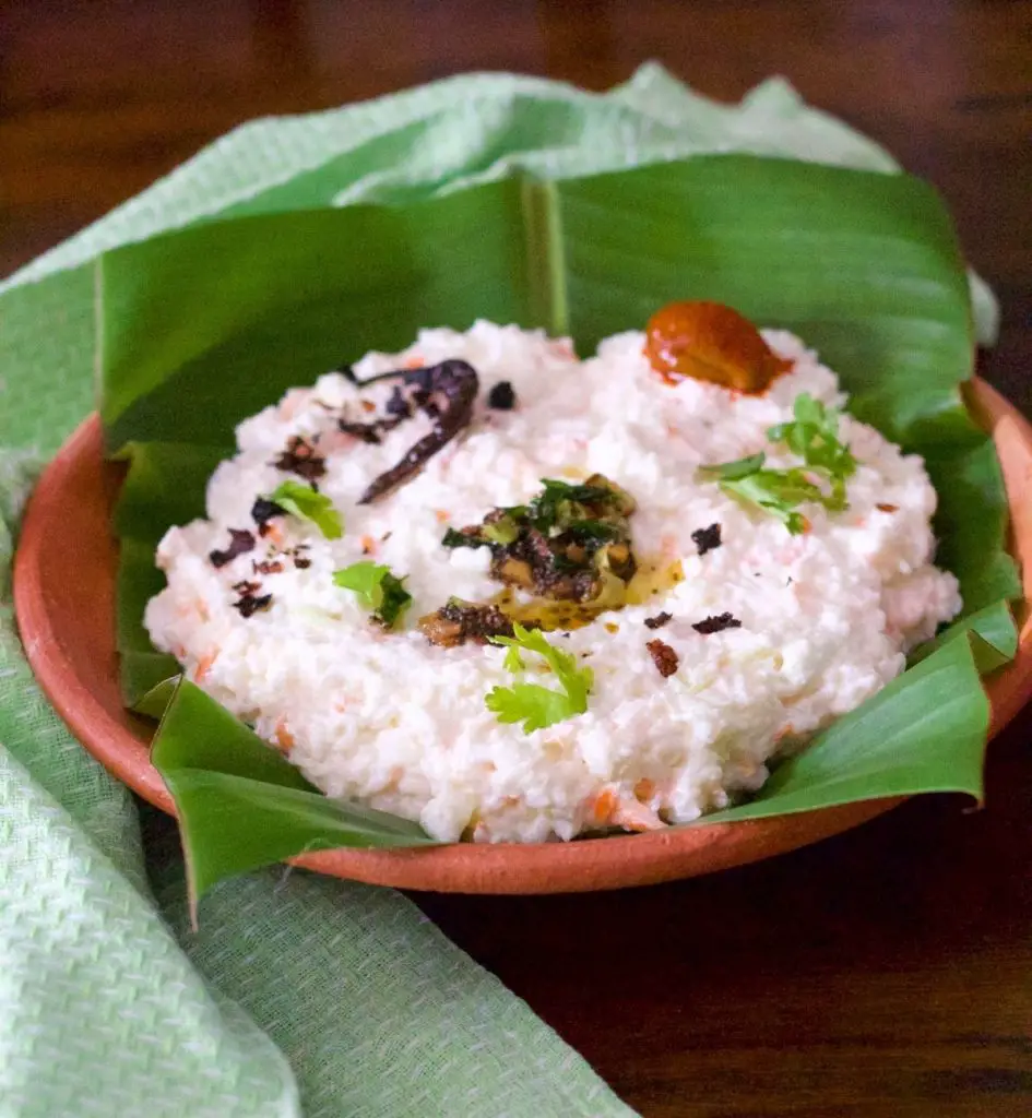 Clay platter filled with rice mixed with yogurt, on a green banana leaf. A pale green napkin on the left. Garnish of green coriander leaves, tempering of mustard seeds and chilies, a small mango pickle seen on top