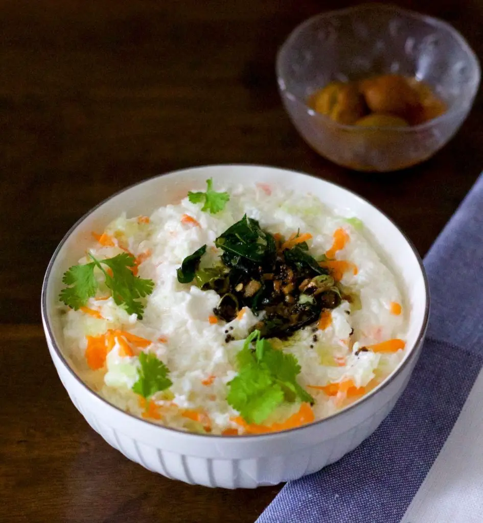 White bowl filled with white curd rice garnished with bright green coriander leaves and orange grated carrot. Tempering of chilies and mustard seeds, curry leaves seen on top. A blue grey and white striped napkin to the right. A bowl of small brownish mango pickles in the background