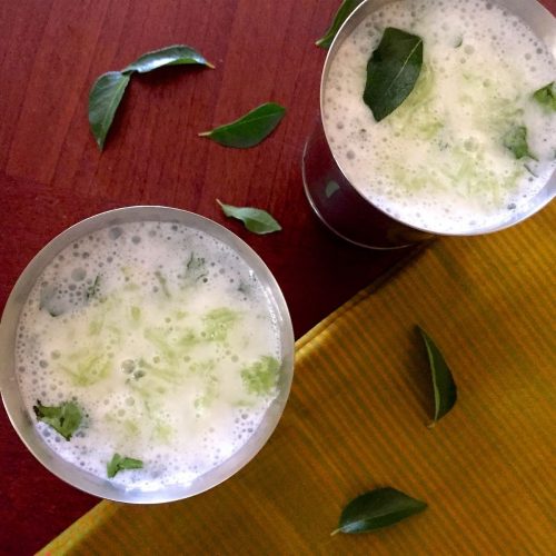 Tall steel glasses of Neer Mor, buttermilk. Green grated cucumber and curry leaves are seen floating on top. A green gold striped saree on the right, curry leaves everywhere, all on a reddish brown background
