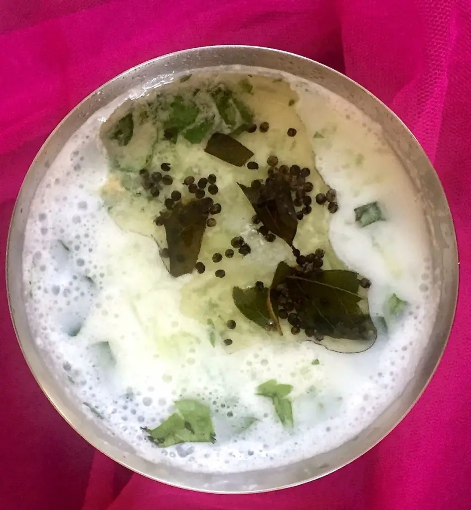 Overhead view of a steel glass of white foamy buttermilk, with cucumber and a tempering of mustard seeds and curry leaves seen floating on top. All on a bright pink fabric background